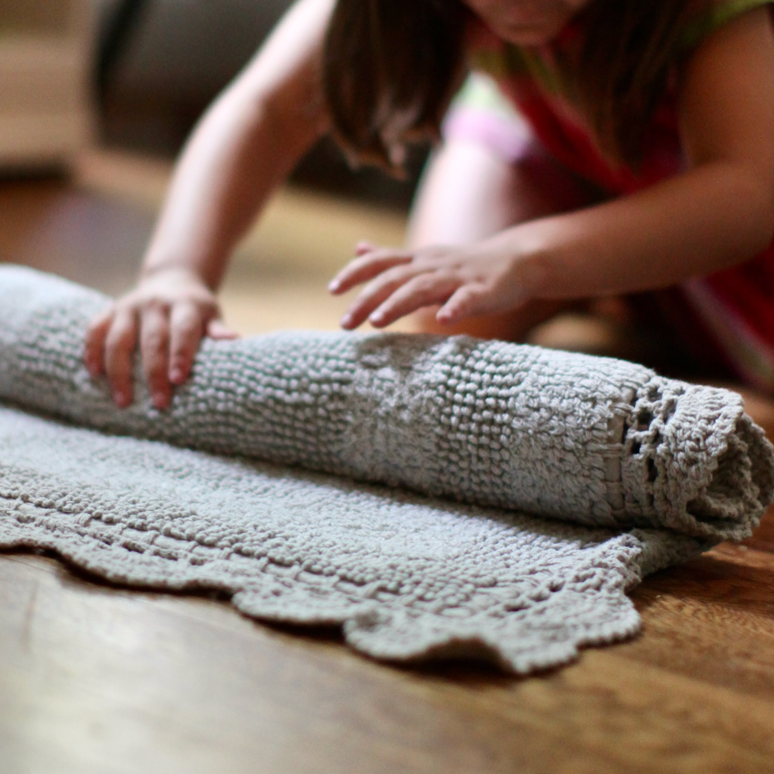 A young child kneeling on a wooden floor, carefully rolling up a crocheted work rug